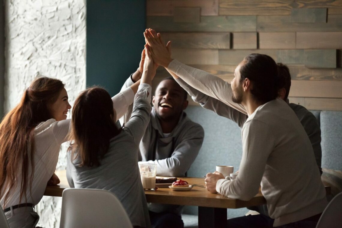 Diverse excited best friends raising hands giving high five together at meeting in cafe, happy multiracial students group promising unity support in good friendly relations hanging in coffee house